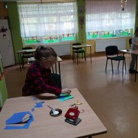 Dziewczynka przykleja wycięte z papieru liście do niebieskiej kartki papieru.