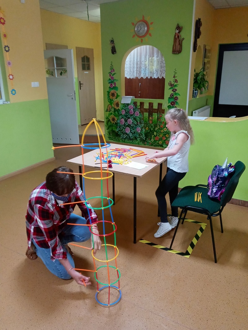 Pani instruktor z Miejskiego Domu Kultury pomaga dziewczynce wykonać konstrukcję z kolorowych patyczków.