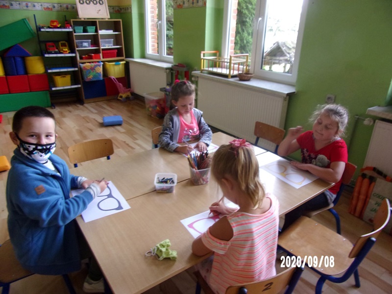 Dzieci siedzące przy stoliku wykonuje zadanie plastyczne, w okularach wydrukowanych na kartce papieru rysują swoje marzenia.
