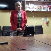 Pani Małgorzata Jarocińska podczas wygłaszania prelekcji, ubrana jest w czerwony żakiet i sukienkę w biało-czarny wzór.