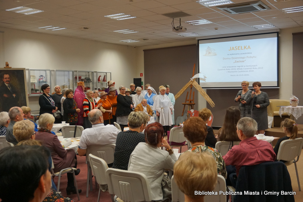 Uczestnicy  Domu Dziennego Pobytu „Zacisze” w Barcinie stoją przed zebraną widownią, dyrektor biblioteki wita zebranych gości