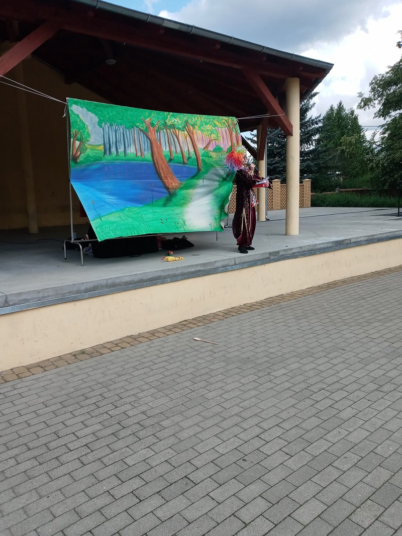 Aktorka wystawiająca teatrzyk przebrana w kolorowy strój oraz kolorową perukę rozpoczyna spotkanie, za aktorką znajduje się tło z namalowanym lasem i jeziorem.
