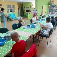 Dzieci biorące udział w zajęciach siedzą przy stołach i słuchają pani prowadzącej.
