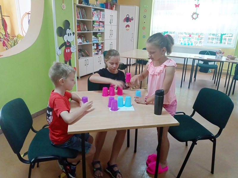 Trójka dzieci siedząca przy stole i układa kolorowe kubeczki według wcześniej otrzymanej instrukcji.