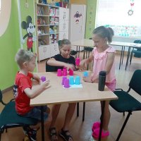 Trójka dzieci siedząca przy stole i układa kolorowe kubeczki według wcześniej otrzymanej instrukcji.