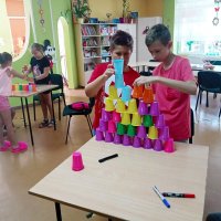 Na zdjęciu widać dwie grupy dzieci, przy stole widocznym na pierwszym planie dwaj chłopcy ustawili piramidę z trzydziestu dwóch kubków, przy stole na drugim planie dwie dzeiwczynki i chłopiec starają się ustawić własną piramidę.