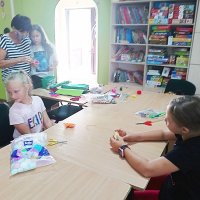 Na pierwszym planie widać te same dwie dziewczynki siedzące naprzeciw siebie, dziewczynka z lewej strony spogląda w bok a dziewczynka z prawej coś do niej mówi, na drugim planie pani pracująca w świetlicy, w której odbywały się zajęcia pomaga innej dziewczynce połączyć kawałki filcu w pąk kwiatu.