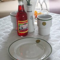 Zastawa stołowa w stylu PRl, widać białe porcelanowe naczynia z zielonym paskiem wokół, butelkę oranżady i papierowy obrus.