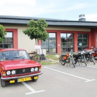 Zdjęcie parkingu przed biblioteką, na którym ustawiono pojazdy z okresu PRL-u, widać czerwonego Fiata 125p zwanego potocznie "dużym fiatem" oraz kilka motocykli.