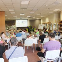 Zdjęcie czytelni biblioteki gdzie odbywało się spotkanie, uczestnicy siedzą przy stolikach i słuchają zaproszonych gości.
