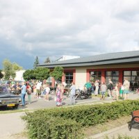 Zdjęcie parkingu przed biblioteką, widać zebranych uczestników spotkania oglądających pojazdy z okresu PRL-u.