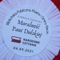 Kotylion przygotowany na Narodowe Czytanie, zawiera napisy Biblioteka Publiczna Miasta i Gminy Barcin ułożony po łuku do góry, Gabriela Zapolska Moralność Pani Dulskiej umieszczony na środku, po niżej tego napisu znajduje się logo akcji, jest to biało-czerwona flaga z dwurzędowym napisem Narodowe Czytanie, na samym dole znajduje się data 04.09.2021r.
