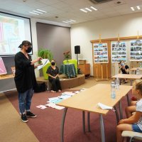Dzieci siedzą przy stolikach, na podłodze rozłożone są kartki papieru, jedna z zaproszonych pań tłumaczy dzieciom, na czym będzie polegało zadania do wykonania.