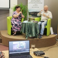 Pani Grażyna Szafraniak ubrana w czarną suknię w różowe kwiaty siedzi na zielonym fotelu ustawionym na scenie, obok niej na takim samym fotelu siedzi Wojciech Przylipiak ubrany w spodnie jeansowe i białą koszulę, pani Grażyna mówi przez mikrofon, pan Wojciech słucha wypowiedzi, popijając wodę, na dole zdjęcia widać laptop na stoliku, dzięki któremu prowadzona jest transmisja na żywo.IMG_0472.JPG