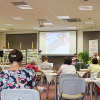 Zdjęcie z tyłu sali, widać uczestników siedzących przy stolikach, stoliki nakryte są serwetkami, na ekranie projektora jest wyświetlany pokaz slajdów złożony ze zdjęć przywiezionych przez Pana Przylipiaka.