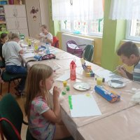 Dzieci siedzą przy nakrytych folią stołach i malują farbami na kartkach papieru korzystając ze słomek.