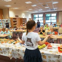 Pani Marzena Żurek stoi za stołami, na których leżą różne owoce i opowiada dzieciom o zdrowym odżywianiu.