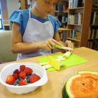Dziewczynka kroi w plasterki banana, na talerzyku ma położonego arbuza a w miseczce leżą truskawki, borówki i maliny.