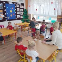 Dzieci siedzą przy stolikach i naklejają czerwone ozdoby na białych sercach wyciętych z papieru.