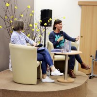Bibliotekarka prowadząca spotkanie wraz z zaproszoną autorką siedzą w fotelach na scenie i rozmawiają.