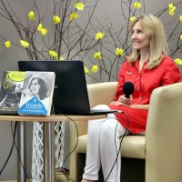 Anna Kamińska ubrana w czerwoną koszulę i białe spodnie, siedzi na beżowym fotelu, przed nią znajduje się laptop i książki.