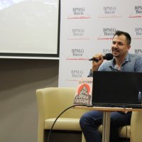 Maciej Piera siedzi w fotelu ustawionym na scenie i mówi do mikrofonu trzymanego w ręku, przed nim na stoliku znajduje się jego książka i laptop.