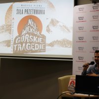 Zdjęcie prowadzącego siedzącego w fotelu na scenie, na ekranie projektora wyświetlane jest zdjęcie książki "Górskie tragedie".