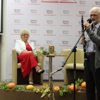 Pani Helena siedzi na fotelu ustawionym na scenie a przed sceną przy mikrofonie stoi Marek Andraszewski.