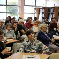 Zdjęcie publiczności, uczestnicy spotkania siedzą przy stolikach i słuchają poezji czytanej przez zaproszonych gości.