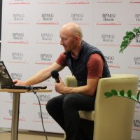 Pan Maciej Stańczak siedzi w fotelu na scenie, w lewej ręce trzyma mikrofon, a prawą przełącza zdjęcia z przygotowanej prezentacji.