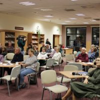 Zdjęcie zebranej publiczności, uczestnicy spotkania siedzą przy stolikach i z uwagą słuchają zaproszonego gościa.