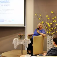 Pani Iwona Kienzler siedzi w beżowym fotelu ustawionym na scenie, wypowiada się do mikrofonu, z lewej strony na rzutniku widać plakat promujący spotkanie, przed sceną widać dyrektor biblioteki siedzącą w fotelu i słuchającą wypowiedzi pani Iwony.
