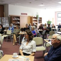 Zdjęcie publiczności, widać dwadzieścia osób, jedna stoi z tyłu sali pozostałe siedzą przy stolikach i słuchają prelegenta.