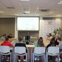 Zdjęcie z tyłu czytelni w bibliotece pokazujące uczniów siedzących przy stołach ustawionych w kształcie podkowy i prowadzącą zajęcia bibliotekarkę siedzącą na krześle przed uczniami.