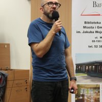 Robert Robb Maciąg stoi przed publicznością i wypowiada się do mikrofonu.