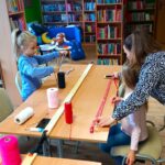 po prawej stronie stołu pracownica biblioteki pomaga przy pomocy miary krawieckiej odmierzyć dziewczynce odpowiednią długość sznurka, a po lewej inna dziewczynka szykuje sobie czarny sznurek do zajęć.
