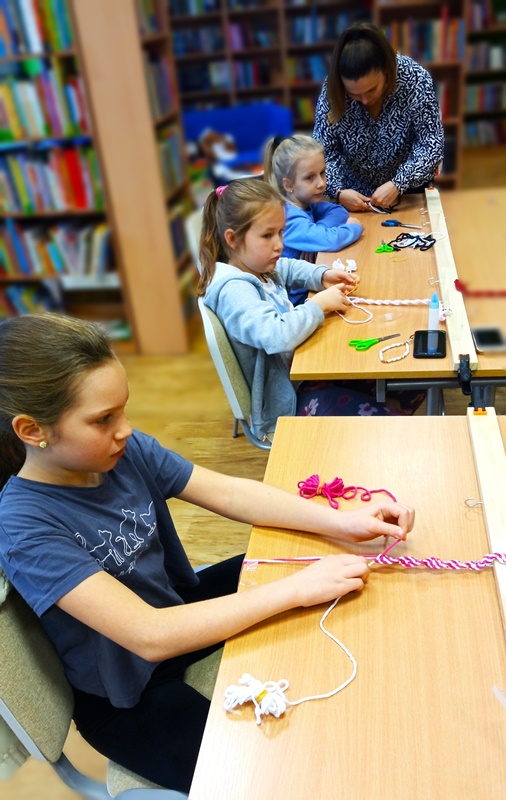 na pierwszym planie dziewczynka siedząca na krześle zaplata różowe sznurki, na drugim planie dwie inne dziewczynki zaplatają sznurki w innych kolorach.