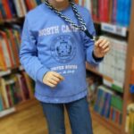 Dziewczynka w niebieskiej bluzie i niebieskich spodniach pokazuje wykonaną na zajęciach smycz do kluczy która zrobiona jest z czarnych i białych sznurków.