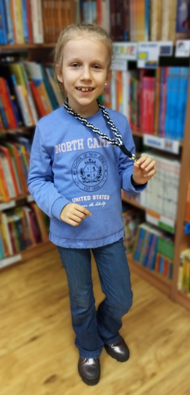Dziewczynka w niebieskiej bluzie i niebieskich spodniach pokazuje wykonaną na zajęciach smycz do kluczy która zrobiona jest z czarnych i białych sznurków.