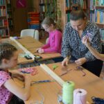 Pracownica biblioteki pomaga dziewczynce podczas zajęć pokazując jej jak należy zaplatać sznurki.