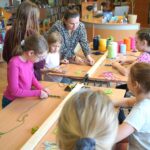 Dzieci pracujące podczas zajęć zaplatają sznurki, pracownica biblioteki pokazuje jak to robić jednej z dziewczynek