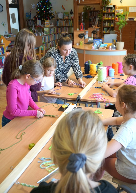 Dzieci pracujące podczas zajęć zaplatają sznurki, pracownica biblioteki pokazuje jak to robić jednej z dziewczynek