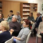 Część publiczności zebranej w bibliotece, ludzi siedzący na krzesłach wokół stolików, przy kawie i ciastkach słuchają wypowiedzi zaproszonego gościa.