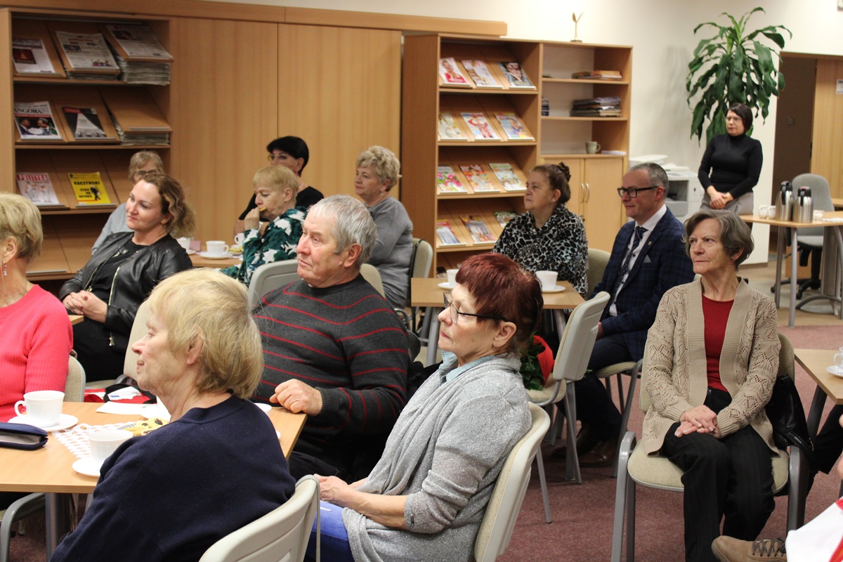 Część publiczności zebranej w bibliotece, ludzi siedzący na krzesłach wokół stolików, przy kawie i ciastkach słuchają wypowiedzi zaproszonego gościa.