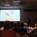 Publiczność zapatrzona w zdjęcie górskiego zbocza prezentowane przez zaproszoną podróżniczkę na ekranie rzutnika, kobieta opowiada o tym co się działo podczas wyprawy w czasie wykonania tej fotografii.