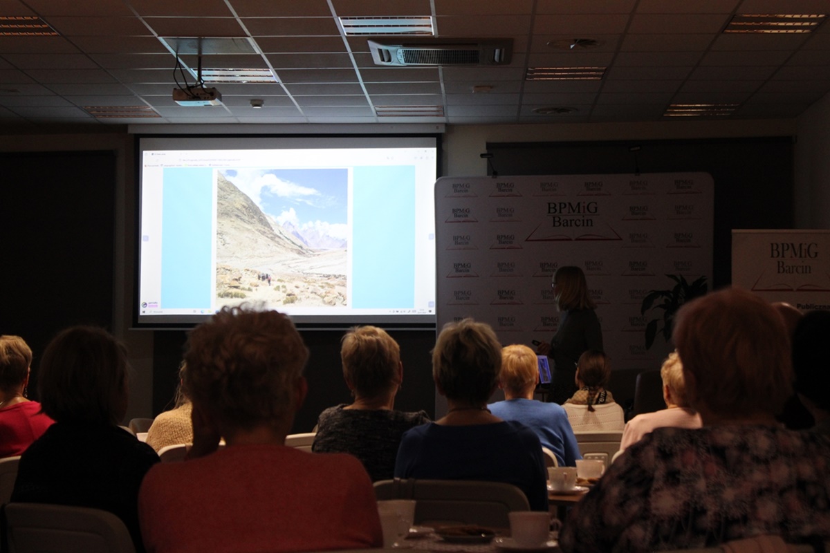 Publiczność zapatrzona w zdjęcie górskiego zbocza prezentowane przez zaproszoną podróżniczkę na ekranie rzutnika, kobieta opowiada o tym co się działo podczas wyprawy w czasie wykonania tej fotografii.