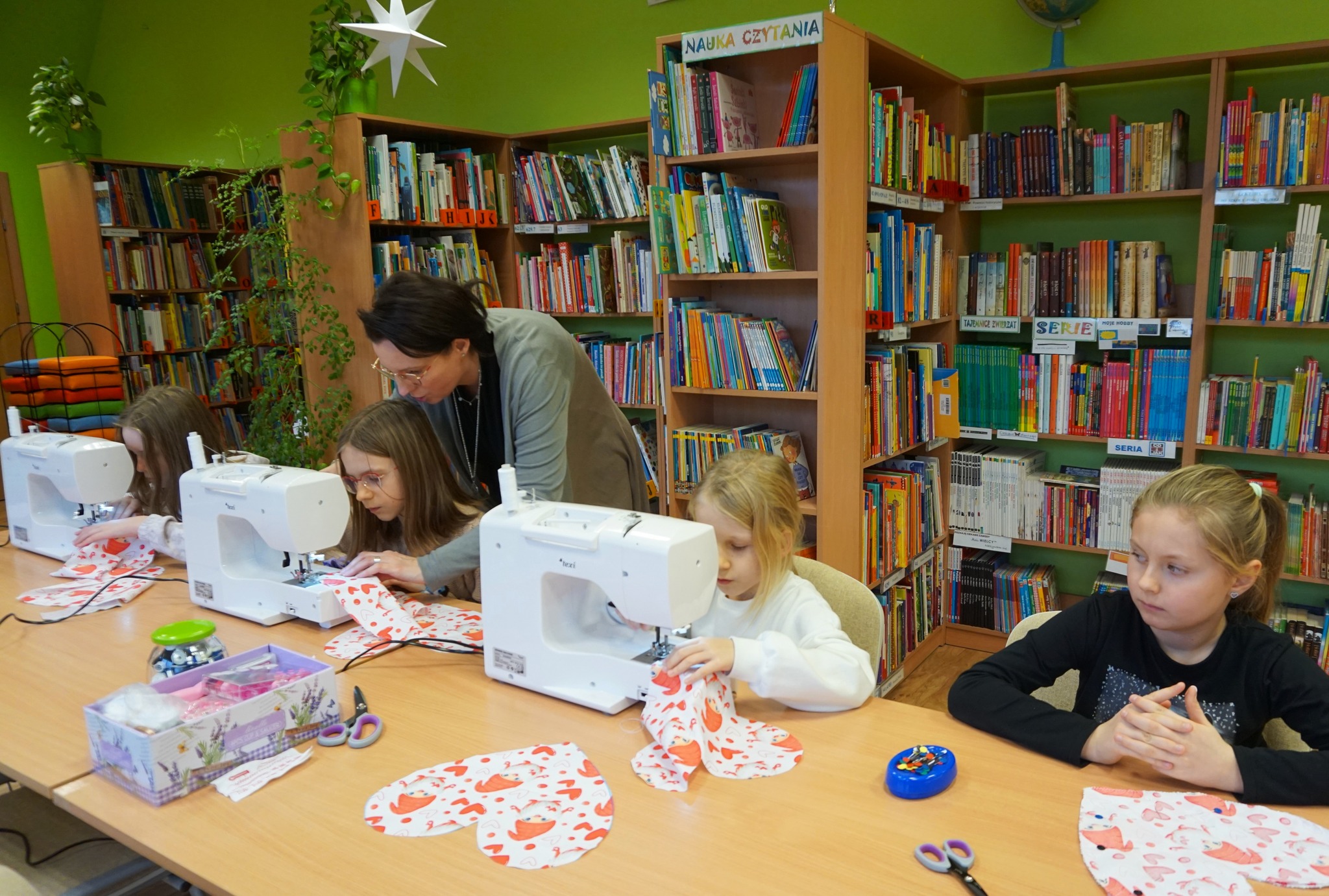 trzy uczestniczki pracują przy maszynach do szycia, jednej z nich pomaga bibliotekarka prowadząca zajęcia, czwarta dziewczynka czeka na swoją kolej