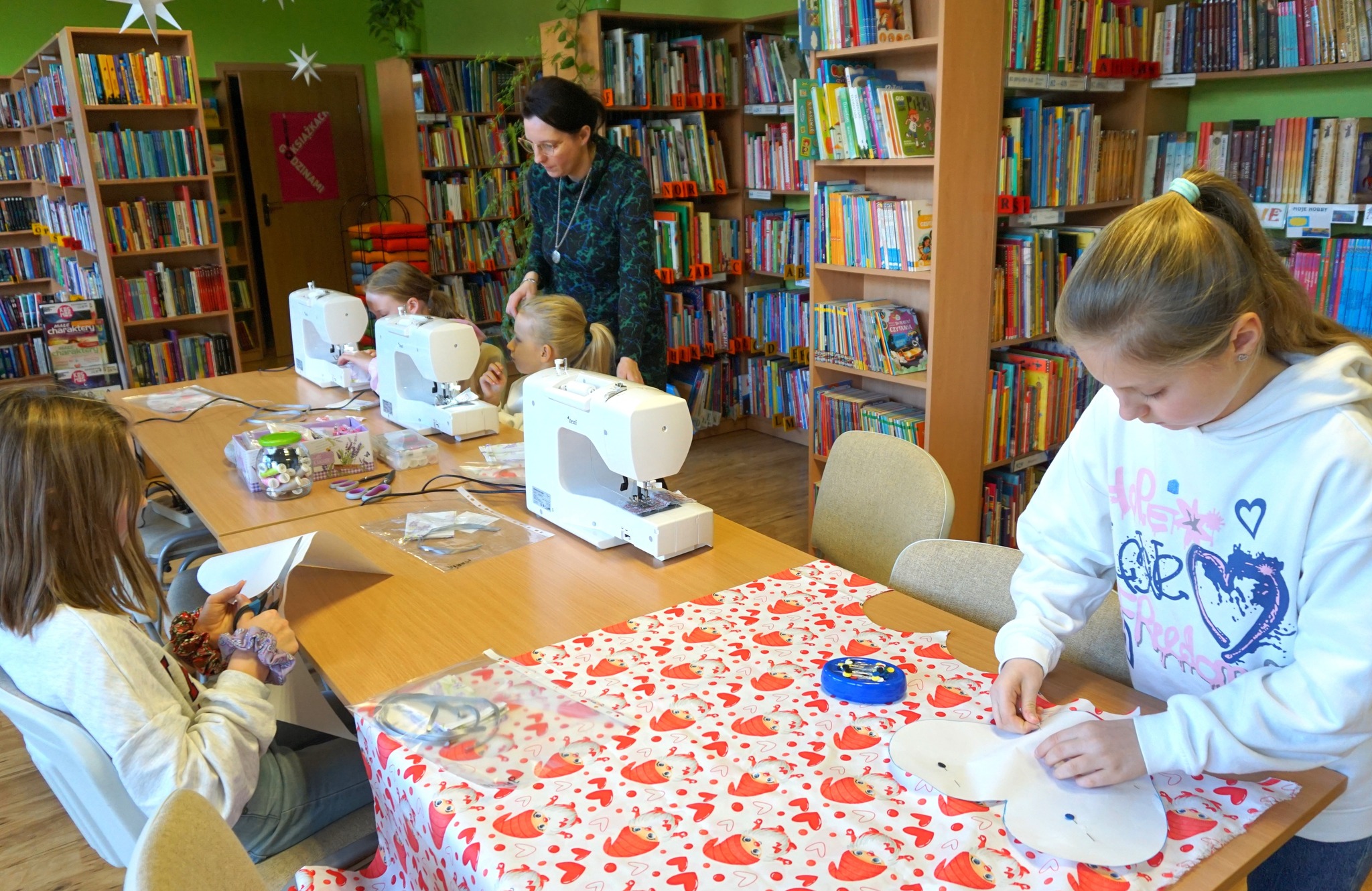 dwie uczestniczki pracują przy maszynach do szycia a dwie kolejne wycina serca z papieru przygotowując się do wycięcia takiego samego kształtu z materiału.