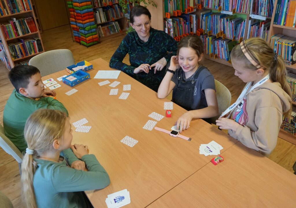 5 uśmiechniętych osób przy stole, czworo dzieci jedna dorosła podczas karcianej gry w której odszukuje się podobne obrazki na tekturowych kartach.