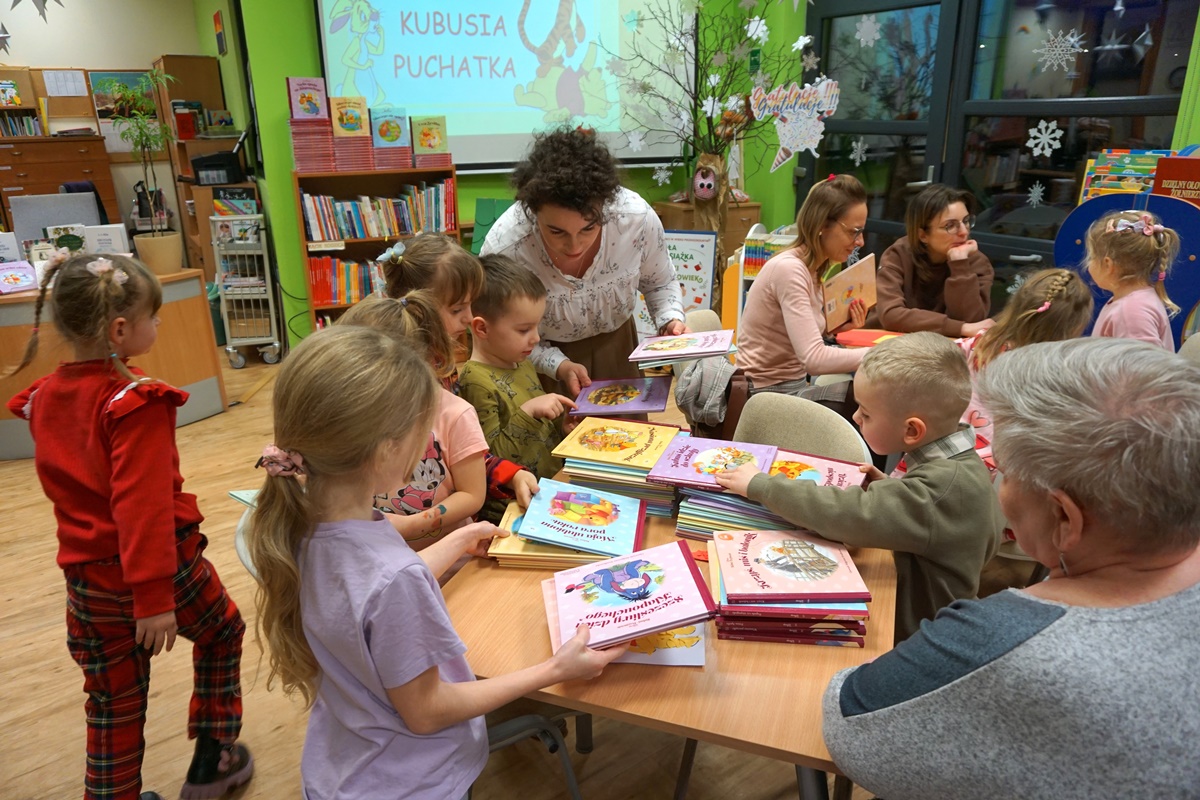 dzieci oglądają książki ułożone na stoliku, jedna z opiekunek opiekunek